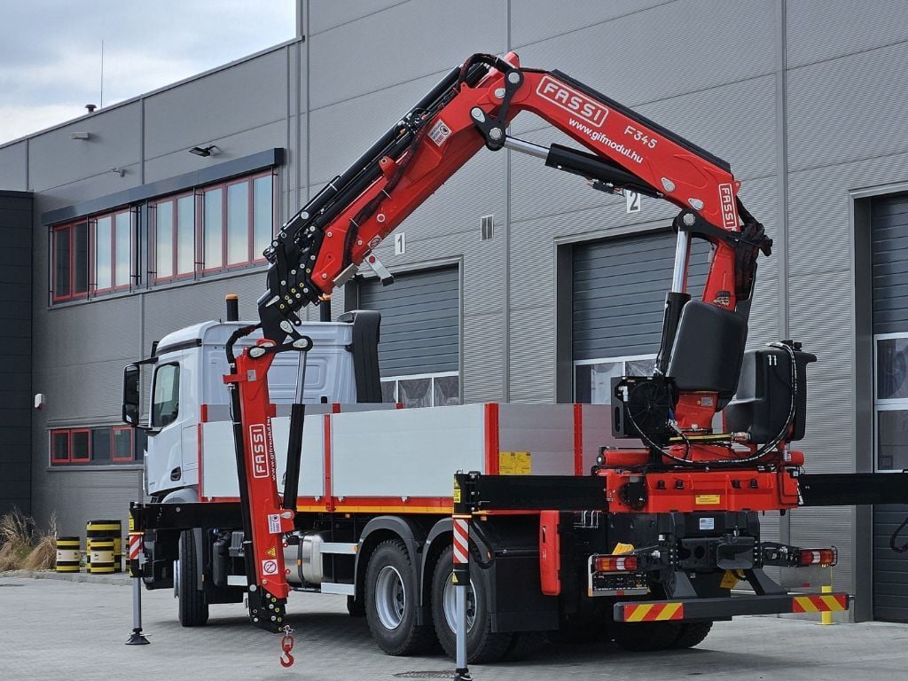Camión equipado con una grúa Fassi con sistema IOC, brazo articulado extendido y estabilizadores desplegados frente a una nave industrial.